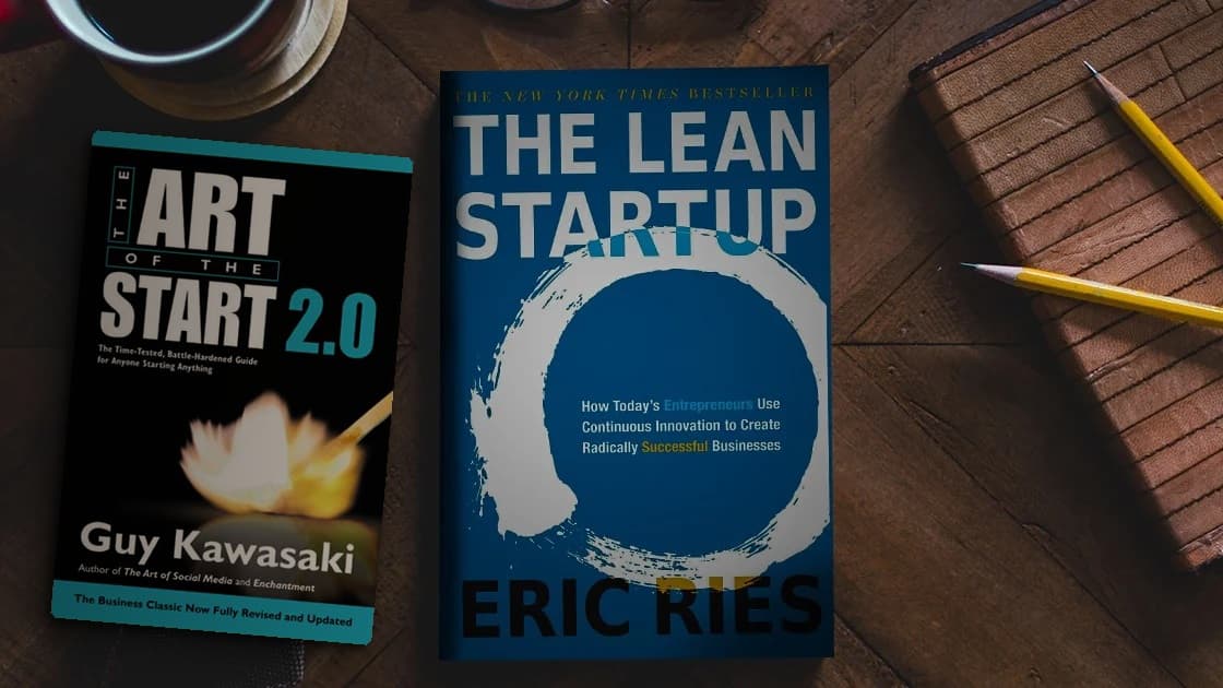 The Art of the Start 2.0 and The Lean Startup lying on a wooden desk next to a notebook, two pencils and a cup of coffee